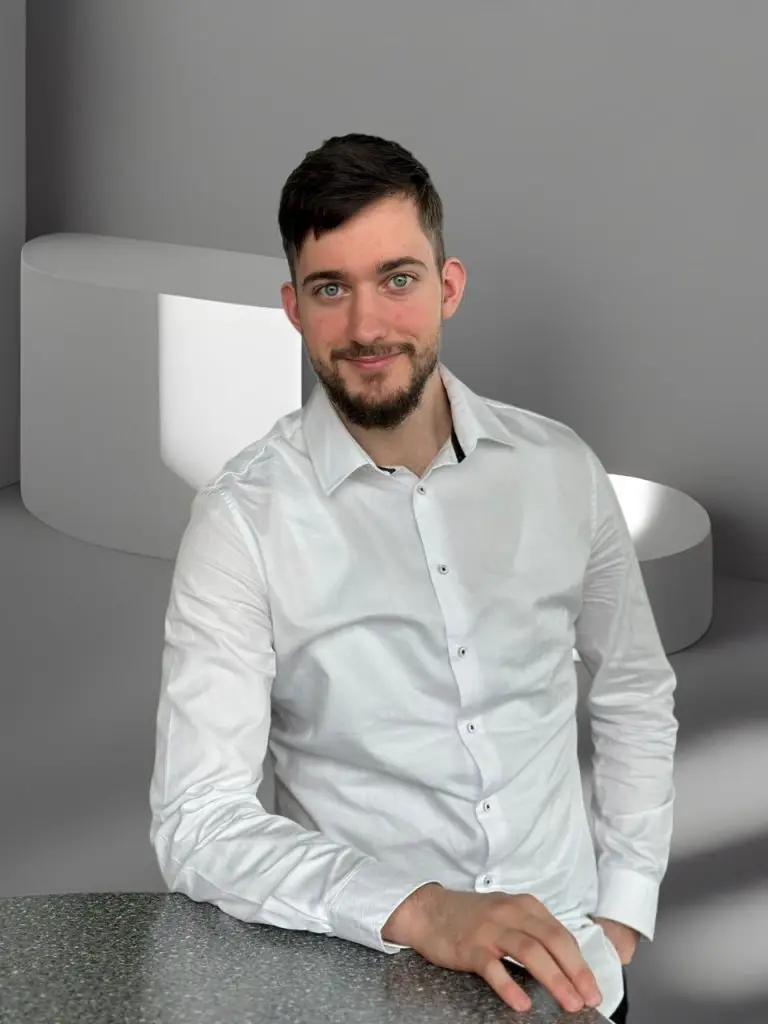 Ein Mann mit dunklem Haar und einem kurzen Bart, der ein weißes Hemd trägt, lehnt sich lächelnd mit seinem Unterarm auf eine graue, gepunktete Tischplatte. Im Hintergrund sind weiße, geometrische Formen in einem Raum mit verschiedenen Grautönen zu sehen.
